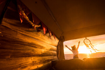 Overjoyed tourist jumping against a golden sunset light viewed from inside a wooden hand made classic van camper. People and travel vanlife lifestyle. Freedom and happiness in summer holiday vacation