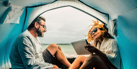 Caucasian adult couple smile and enjoy leisure inside a tent in free camping at the beach with sea view. Concept of alternative tourist and travel vacation. Woman using tablet and connection
