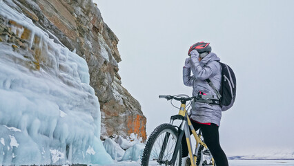 Woman is riding bicycle near the ice grotto. The rock with ice caves and icicles. Girl is dressed...