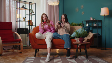 Worried funny girls friends siblings enthusiastically playing racing video games on smartphone. Happy two female women couple family on sofa at home, using smartphone gadget app with drive simulator