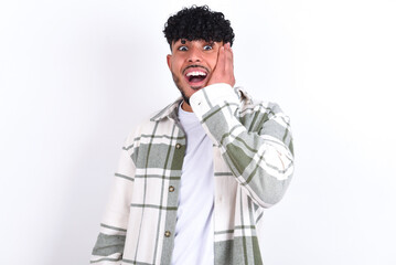 Shocked, astonished young arab man with curly hair wearing overshirt  looking surprised in full disbelief wide open mouth with hand near face. Positive emotion facial expression body language.