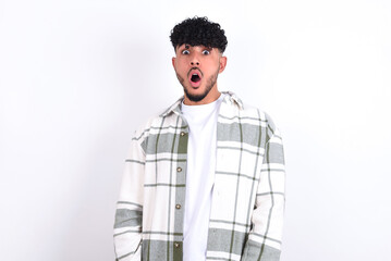 Expressive facial expressions. Shocked stupefied young arab man with curly hair wearing overshirt over white background, keeps jaw dropped feels stunned from what he sees aside.