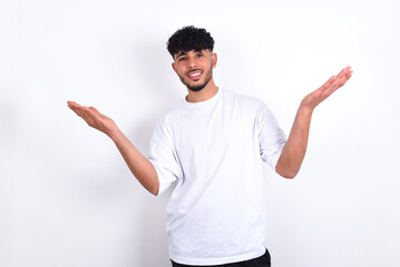 Cheerful young arab man with curly hair wearing white t-shirt over white background  making a welcome gesture raising arms over head.