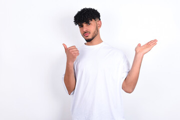 Happy cheerful young arab man with curly hair wearing white t-shirt over white background  showing thumb up and pointing with the other hand. I recommend this.