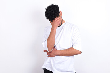 young arab man with curly hair wearing white t-shirt over white background  making facepalm gesture...