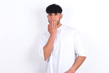 Oh! I think I said it! Close up portrait young arab man with curly hair wearing white t-shirt over white background  cover open mouth by hand palm, look at camera with big eyes.