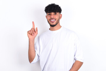 Pleasant looking young arab man with curly hair wearing white t-shirt over white background  has clever expression, raises one finger, remembers himself not to forget tell important thing.