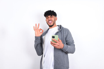 Happy young arab man with curly hair wearing casual clothes over white background sending a message on his smartphone or taking a selfie  and making ok sign with his hand. - obrazy, fototapety, plakaty