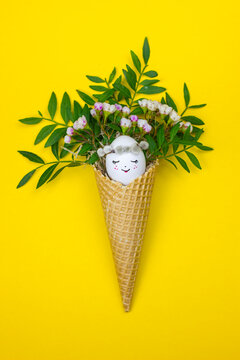 Easter egg with cute face in the waffle cone with spring flowers on yellow background.