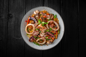 Soba, udon, asian wheat noodles with seafood: shrimps, mussels, squid and vegetables: broccoli, carrots, sweet peppers, zucchini on a black background. Asian style. Japanese food