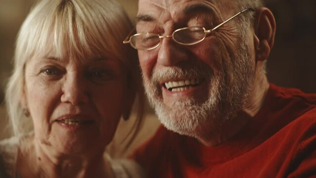 Senior couple smiling and talking with online relatives while making video call via cellphone on weekend day at home