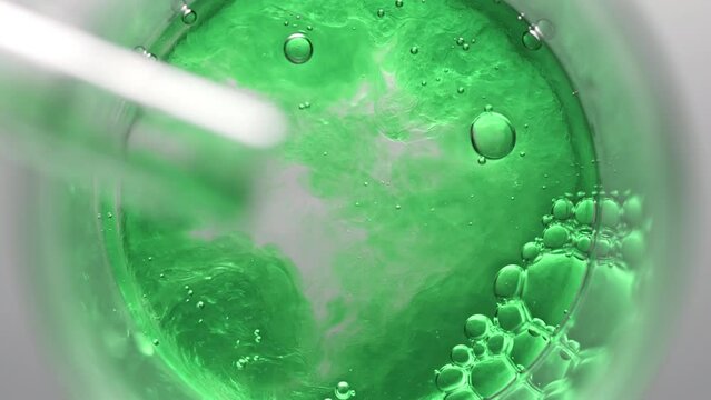 Green liquid pouring into beaker with water and glycerin creates different sized bursting bubbles and ripples on its surface on light grey background | Abstract body care cosmetics mixing concept