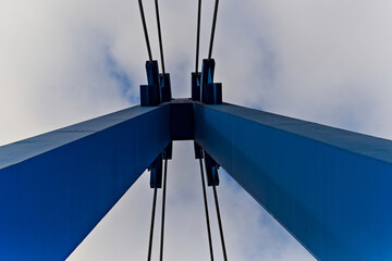 Pylon wiszącego mostu w kolorze niebieskim ze stalowymi linami nośnymi na tle szarego , zachmurzonego , zimowego nieba .