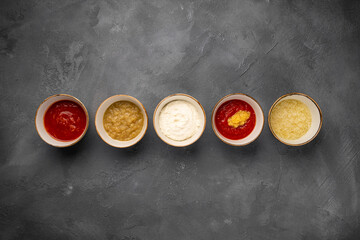 Set of different sauces in bowls and ingredients on gray rustic concrete background, top view. Tomato ketchup, mayonnaise, guacamole, mustard, soy sauce, pesto, cheese sauce - assortment of dips