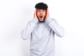 young arab man with curly hair wearing sport sweatshirt
over white background with scared expression, keeps hands on head, jaw dropped, has terrific expression. Omg concept