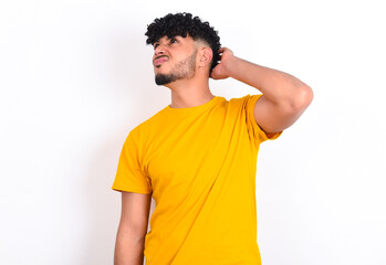 young arab man wearing yellow T-shirt over white background confuse and wonder about question. Uncertain with doubt, thinking with hand on head. Pensive concept.