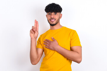 young arab man wearing yellow T-shirt over white background smiling swearing with hand on chest and...