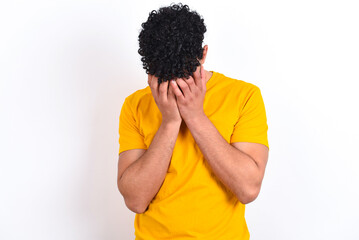 young arab man wearing yellow T-shirt over white background covering her face with her hands, being...