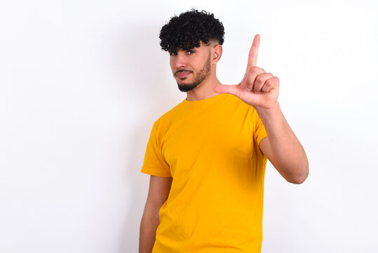 young arab man wearing yellow T-shirt over white background making fun of people with fingers on forehead doing loser gesture mocking and insulting.