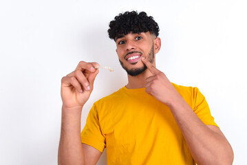 young arab man wearing yellow T-shirt over white background holding an invisible aligner and pointing to her perfect straight teeth. Dental healthcare and confidence concept. - obrazy, fototapety, plakaty