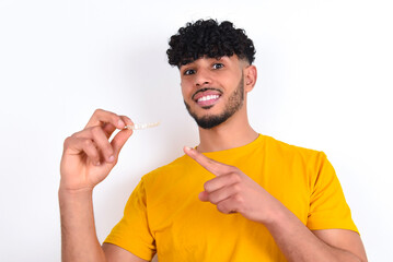 young arab man wearing yellow T-shirt over white background holding an invisible aligner and...