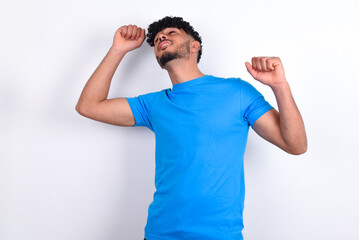 Photo of upbeat young arab man with curly hair wearing blue t-shirt over white background has fun and dances carefree wear being in perfect mood makes movements. Spends free time on disco party