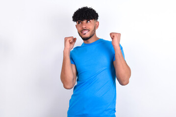 young arab man with curly hair wearing blue t-shirt over white background clenches fists and awaits...