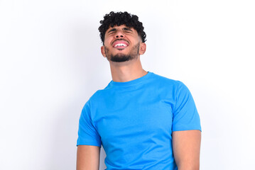 Fototapeta premium Positive young arab man with curly hair wearing blue t-shirt over white background with overjoyed expression closes eyes and laughs shows white perfect teeth