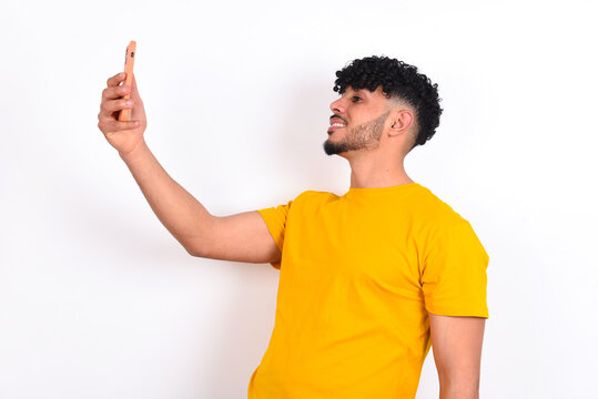 young arab man wearing yellow T-shirt over white background smiling and taking a selfie ready to post it on her social media.