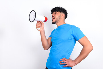 Funny young arab man with curly hair wearing blue t-shirt over white background People sincere emotions lifestyle concept. Mock up copy space. Screaming in megaphone.