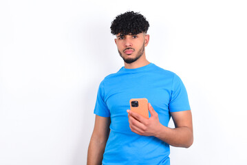 Upset dissatisfied young arab man with curly hair wearing blue t-shirt over white background uses mobile software application and surfs information in internet, holds modern mobile hand