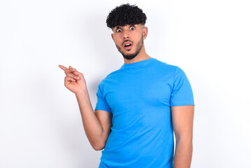 Emotive young arab man with curly hair wearing blue t-shirt over white keeps jaw dropped from shock demonstrates amazing promo points right on blank space demonstrates big shopping sale. Advertisement