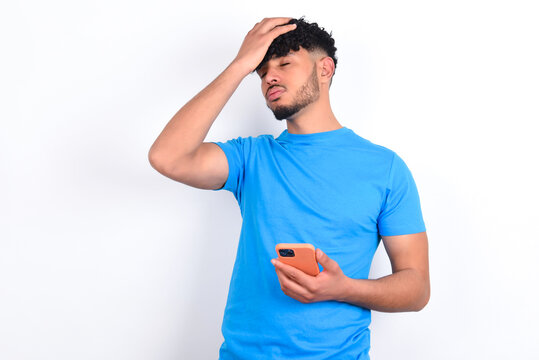 Upset Depressed Young Arab Man With Curly Hair Wearing Blue T-shirt Over White Background Makes Face Palm As Forgot About Something Important Holds Mobile Phone Expresses Sorrow And Regret Blames