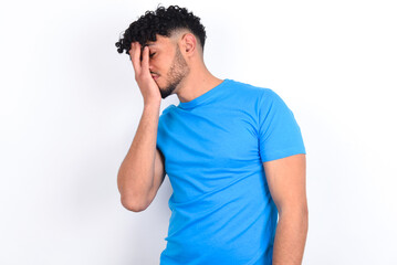 young arab man with curly hair wearing blue t-shirt over white background with sad expression covering face with hands while crying. Depression concept.