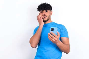 Afraid funny young arab man with curly hair wearing blue t-shirt over white background holding telephone and bitting nails