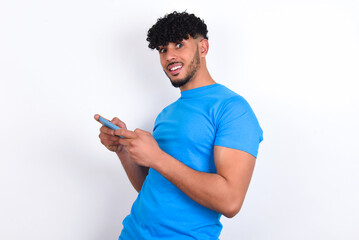 Nice addicted cheerful young arab man with curly hair wearing blue t-shirt over white background using gadget playing network game