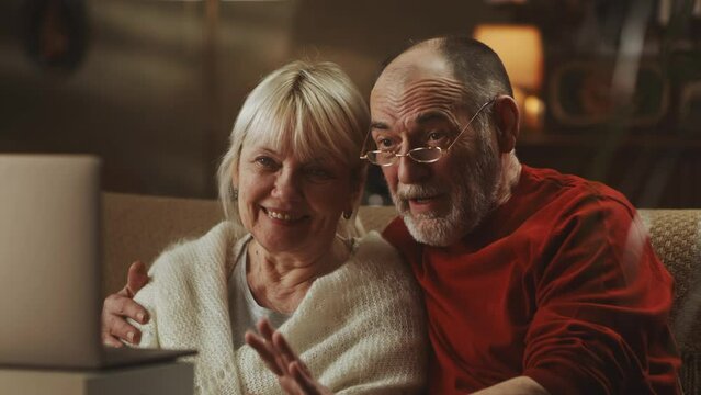 Glad elderly man embracing smiling wife and talking with relatives while making video call via netbook at home