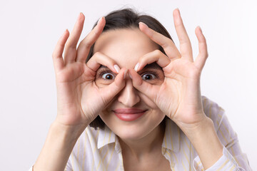 Close up portrait of attractive quirky young woman making binoculars with hands showing ok gesture on white studio background.