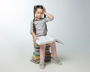 little girl with braided pigtails sits on a large stack of books and presses her hand to her head