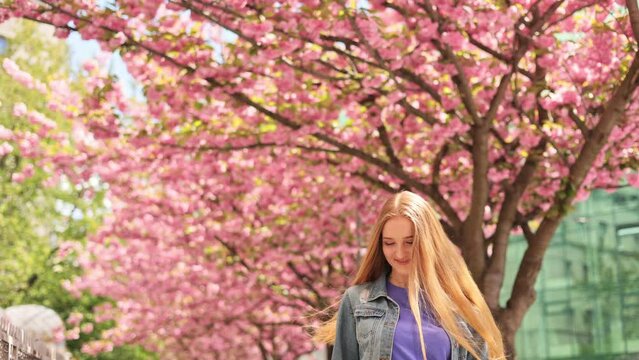 Beautiful young ukrainian girl feels free and happy look around and smiling on blurred background in the city street. Sakura tree. Caucasian woman with long hair walking around Cherry blossom trees.