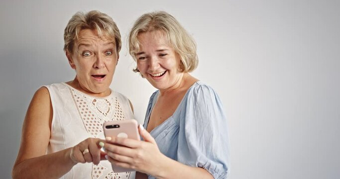 Two shocked casual young female friends with mobile phone against white background
