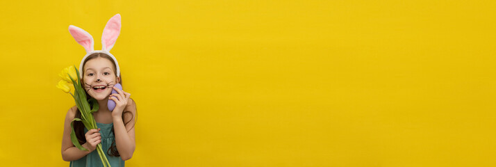 Little happy girl with long dark hair in a headband with rabbit ears holds colorful purple Easter eggs and yellow tulips near her face and looks at the camera on a yellow background, easter card