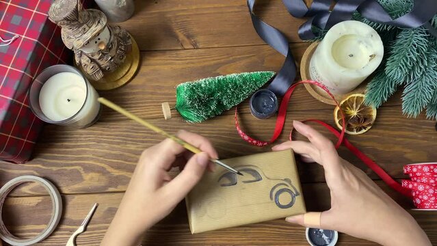 Human's hands draw car with brush and paint on gift box. New Year preparations. Surprise in craft wrapper, decorative Christmas tree, candles, ribbons and snowman figure on wooden table.