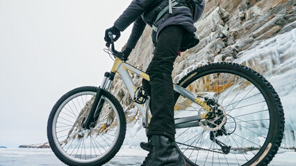 Fototapeta na wymiar Man is riding bicycle near ice grotto. Rock with ice caves icicles. Teenage is dressed in black down jacket, cycling backpack, helmet. Tires on covered with special spikes. Traveler boy is ride cycle.