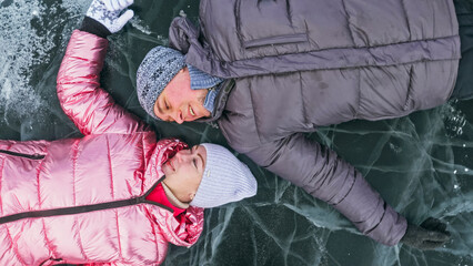 Couple has fun winter walk against background of ice of frozen lake. Lovers lie on clear ice with cracks have fun kiss and hug. View from above. Happy people on snow covered ice. Honeymoon love story.