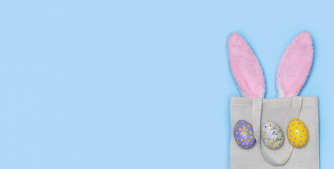 Banner. Pink Easter bunny ears sticking out of Reusable textile bag with Painted eggs on a blue background. Happy Easter minimal concept. An Easter card with a copy of the place for the text. Flat lay
