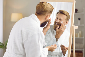 Smiling young man look in mirror apply moisturizing face cream take care of body hygiene. Happy millennial guy use facial lotion or balm for smooth skin. Skincare and male beauty procedure concept.