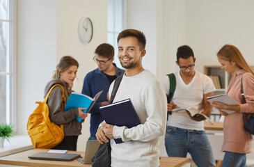 Fototapeta Portrait of a happy male college or university student in the classroom. Handsome young man holding a textbook and smiling at the camera while his classmates are studying new books in the background obraz