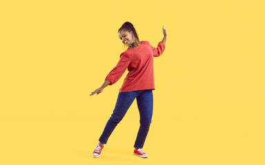 Happy young African American woman on yellow studio background dancing in casual clothes. Smiling millennial teen black girl have fun show dancer moves. Hobby and entertainment.