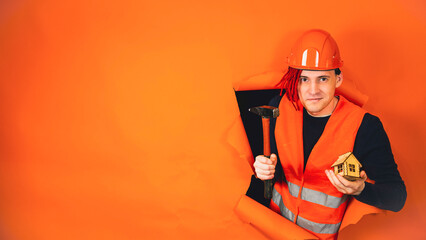 Male construction worker in overalls holds wooden house and sticks out of hole of orange background. Man in hard hat with toy house and hammer. Concept of purchasing new apartment and roof overhead.